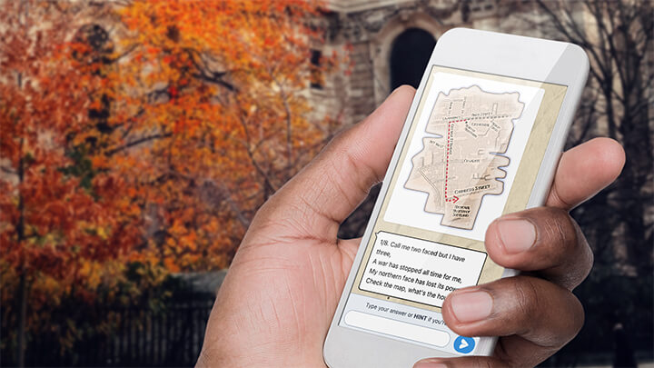 A hand holding a phone playing Treasure Hunt Cambridge with a tree with orange leaves behind.